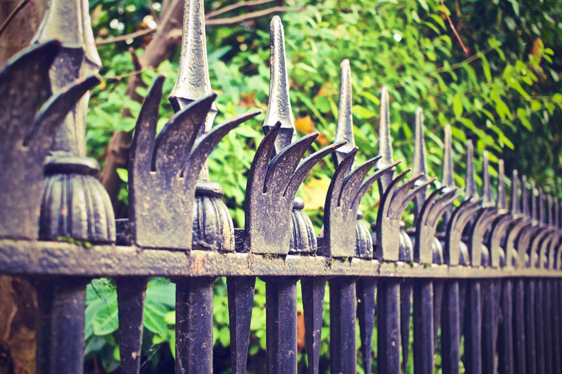 Close-up of a metal fence top rail lined with pointed spike finials in front of green shrubs