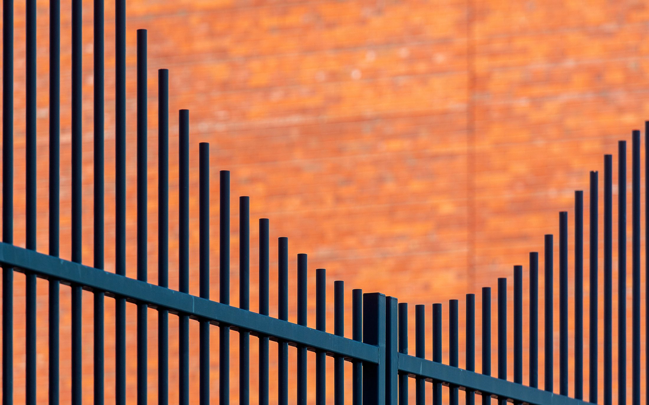Black flat-top metal picket fence stepping along a sloped run in front of an orange brick wall