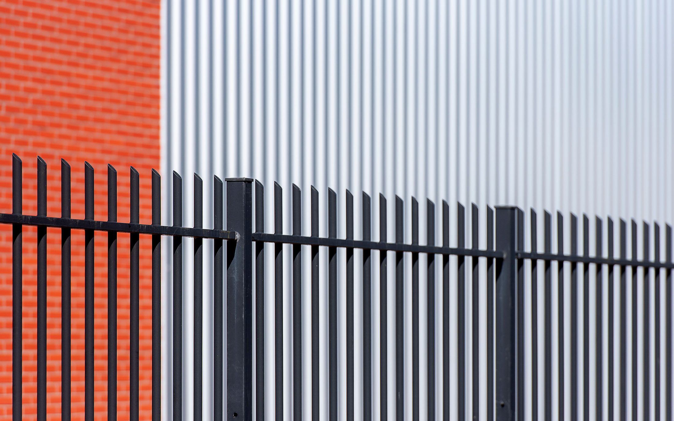 Black metal picket fence panels and posts running in front of a red brick and corrugated metal wall