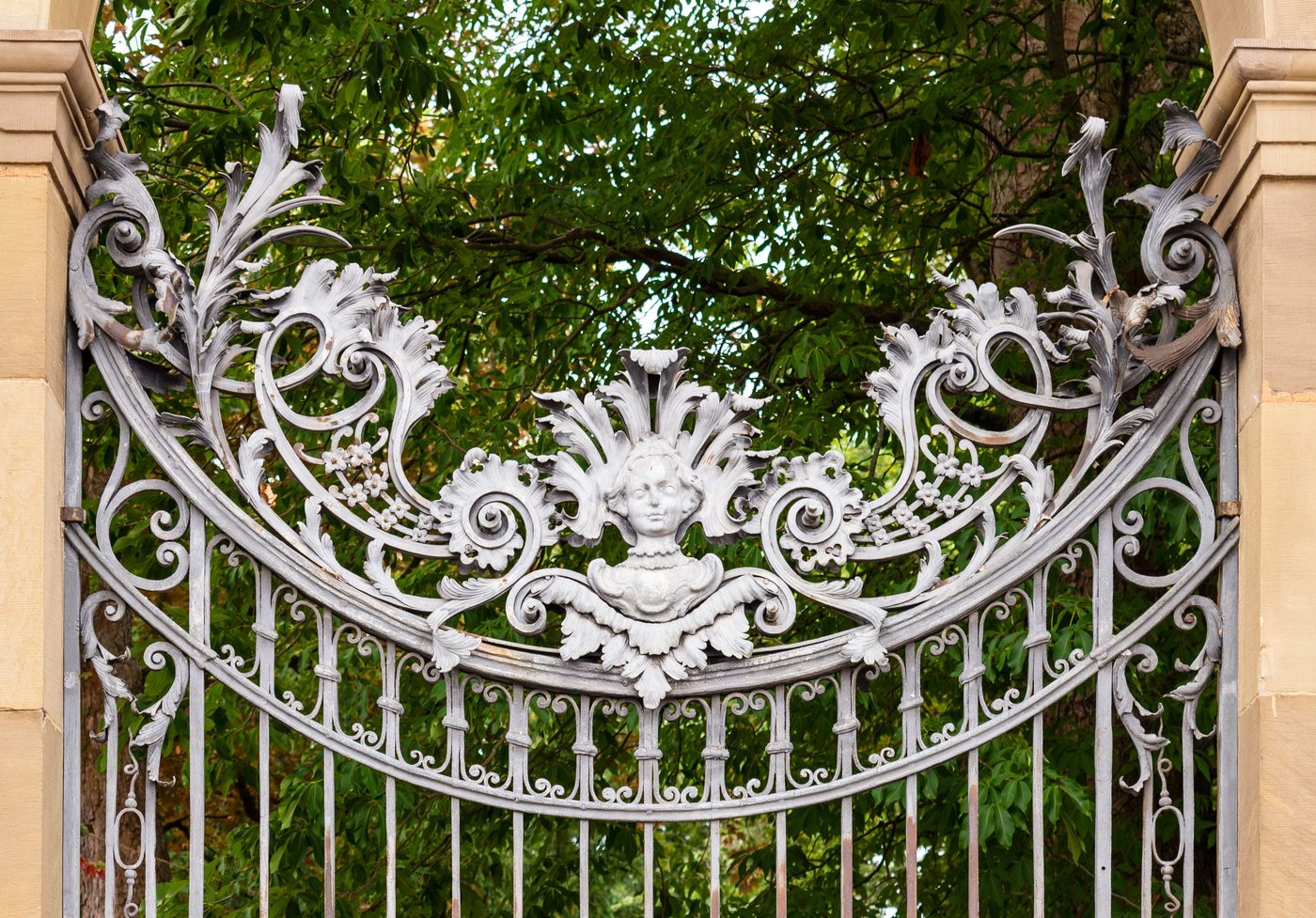 Silver-painted wrought iron gate top with leaf scrolls and a mask between sandstone posts