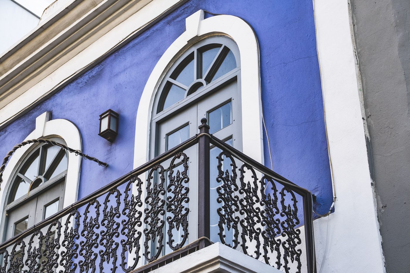 Black ornate iron balcony railing on a blue facade with arched white windows