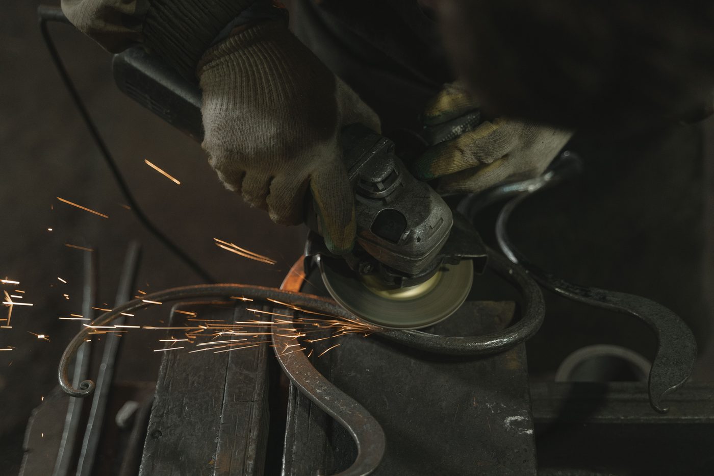 Gloved hands using an angle grinder on curved metal scroll pieces, throwing sparks