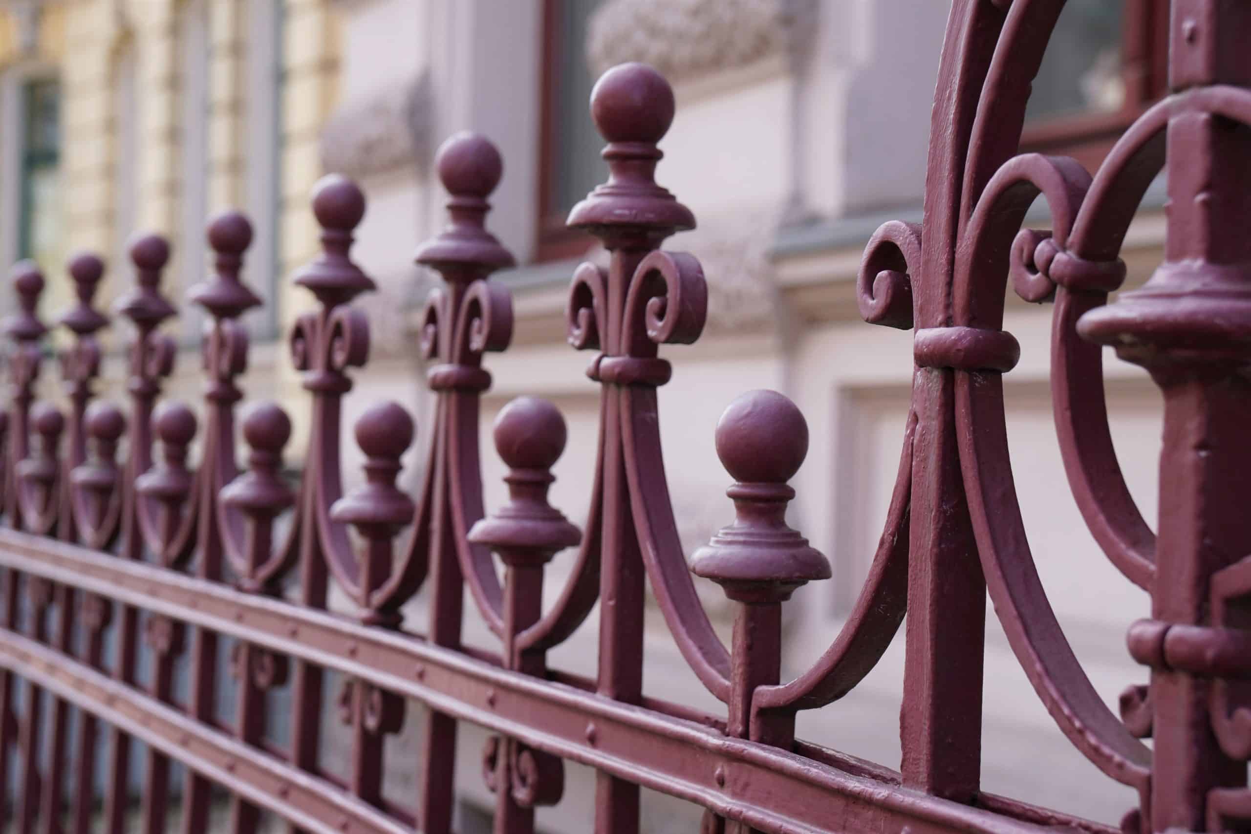 Ornate red iron railing with decorative scrollwork and finials along an urban street
