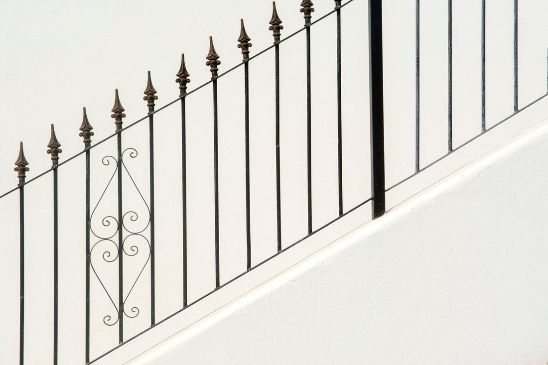 Close-up of a black wrought iron railing with spear finials and a scroll insert against a white wall