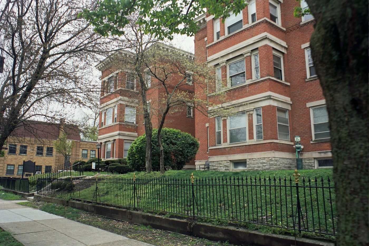 Black iron picket fence along a lawn and sidewalk in front of red brick apartment buildings